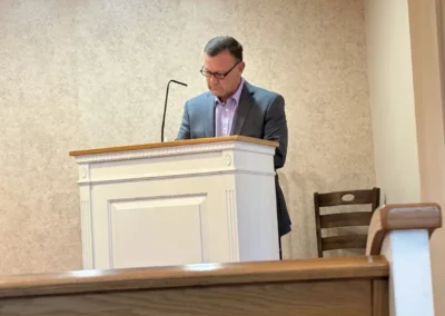 A speaker stands behind a wooden podium in a simple room, with a chair visible in the background