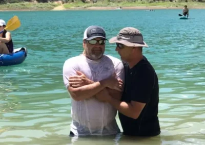 Two men stand in a shallow lake, one with wet clothing. A kayak and paddleboard are visible in the background under a sunny sky